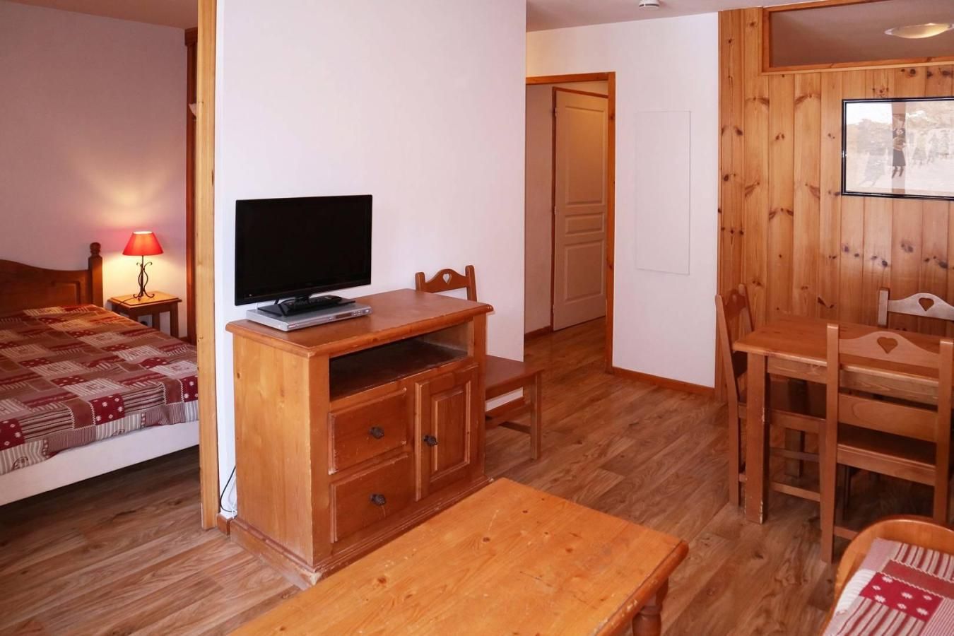 A bedroom with a bed, a TV on a wooden stand, a bedside table with a lamp, and a wooden floor.