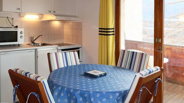 A compact kitchen with a microwave, stove, and sink. There is a dining table with striped chairs and a blue tablecloth. A yellow curtain is partially visible, and a wooden door leads to an outdoor area. - 50m from resort center, 4th floor, balcony, 22m², Les Orres (Les Orres)