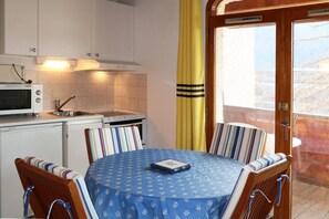 Eine kleine Küche mit Mikrowelle, Herd und Spüle. Ein Esstisch mit gestreiften Stühlen und einer blauen Tischdecke. Ein gelber Vorhang ist teilweise zu sehen, und eine Holztür führt in einen Außenbereich.