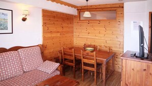 Una habitación acogedora con pared de madera, un sofá, una mesa de comedor con sillas, una mesita con un tazón y un televisor sobre un mueble, además de una lámpara en la pared.