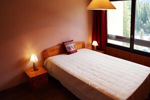 Een opgemaakt bed met een witte dekbedovertrek en een rood kussen, twee nachtlampjes, een houten nachtkastje en een raam met uitzicht op bomen.