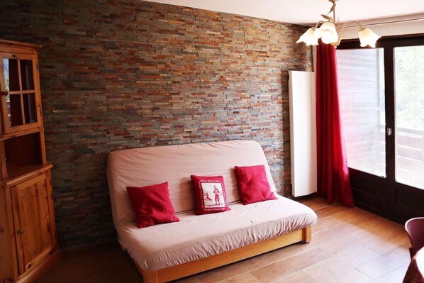A room with a brick wall, a wooden cabinet, a sofa with red cushions, and a window with red curtains.