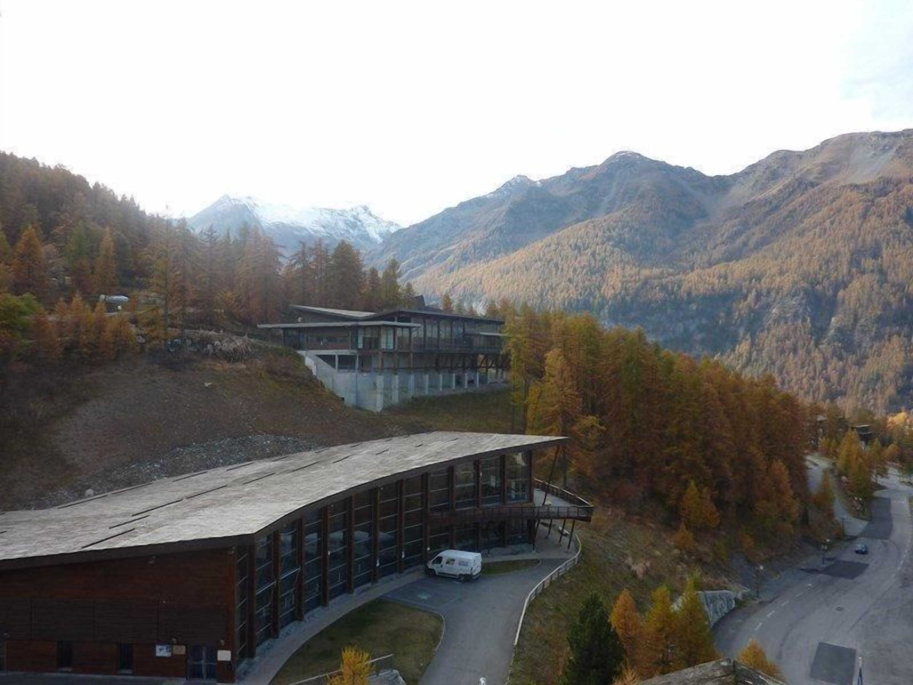 Een modern gebouw met een golvend dak, omgeven door een bergachtig landschap met herfstkleuren.