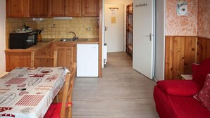 A compact kitchen with wooden cabinets, a microwave, and a sink. There is a small refrigerator and a dining table with a red chair. A hallway leads to a bedroom with a red bed. - 15m from ski slopes, 5th floor, view valley, balcony, 22m², Les Orres (Les Orres)