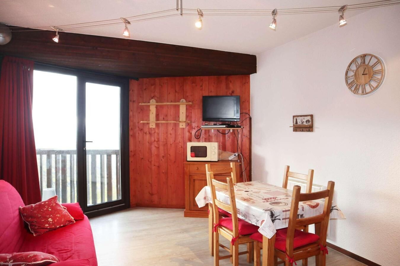 A cozy living room with a red sofa, a dining table with chairs, a TV mounted on a wooden wall, and a wooden ceiling.