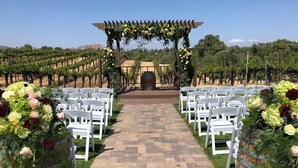 Un viñedo con un sendero que lleva a una pérgola decorada con flores, flanqueada por sillas blancas.