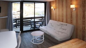 Ein Zimmer mit einer Couch, einem kleinen runden Tisch und einem Esstisch. Eine Glastür führt zu einem Außenbereich mit Stühlen.