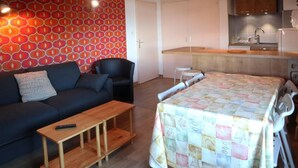 A compact living space with a sofa, a small table, a bed with a quilt, a kitchen area, and a patterned wall.