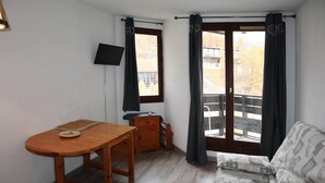 Een kleine kamer met een houten tafel, een bank, een aan de muur bevestigde televisie en een raam met gordijnen.