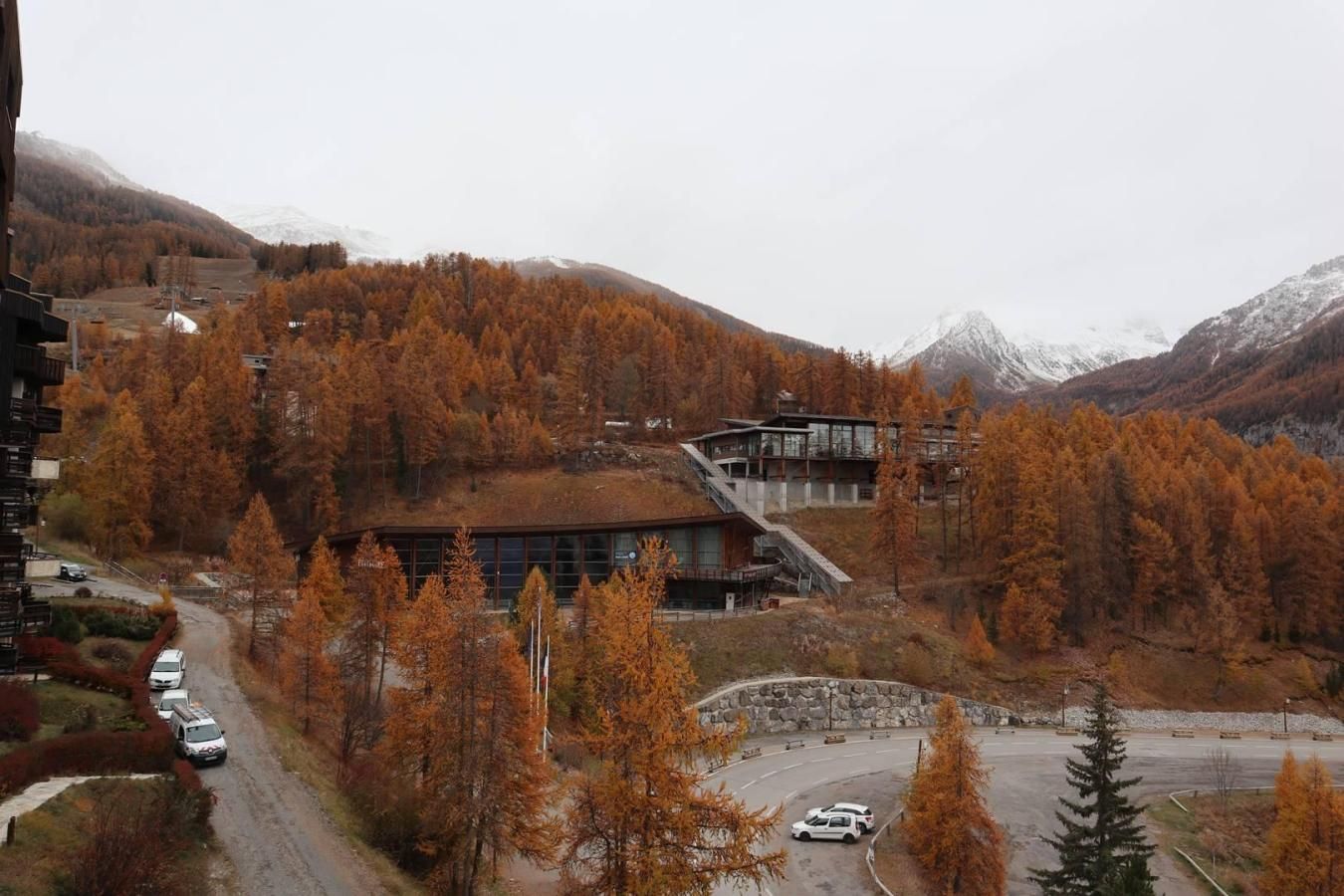 A modern building with a glass facade, surrounded by autumn-colored trees and a mountainous backdrop.