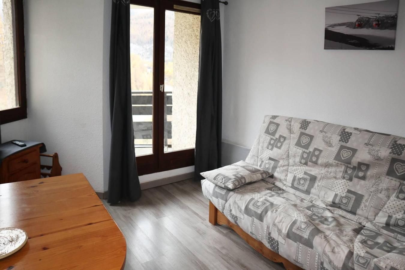 Una habitación con una mesa de madera, un sofá con cojines estampados, una ventana con cortinas y un cuadro colgado en la pared.