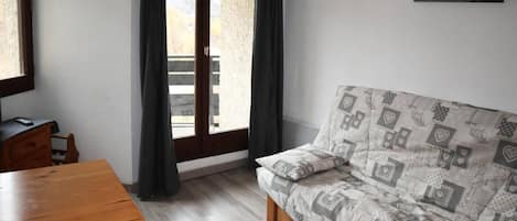 Una habitación con una mesa de madera, un sofá con cojines estampados, una ventana con cortinas y un cuadro colgado en la pared.