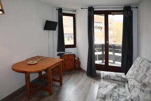 A room with a wooden table, a sofa, a TV mounted on the wall, and a balcony with curtains. - 50m from ski slopes, 4th floor, balcony, 26m², Les Orres (Les Orres)
