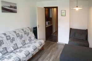 A room with a patterned sofa, a dark armchair, a hanging light fixture, and a door leading to a hallway.