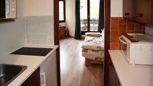 Een compacte keuken met een spoelbak, magnetron en een kleine koelkast. Een bed met een gebloemd plaid is zichtbaar door een open deur.
