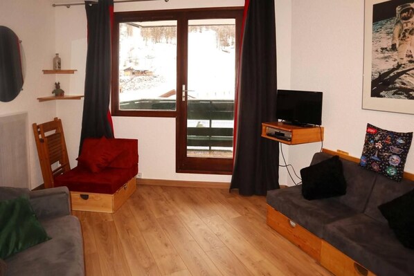 A room with a wooden floor, a red chair, a grey sofa, a television on a wooden stand, and a window with curtains.