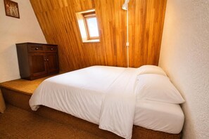 Une chambre avec un plafond en pente, une petite fenêtre, un lit avec une literie blanche et une commode en bois.