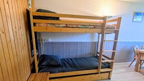 Ein Stockbett mit Holzleiter, ein kleiner Holztisch und ein Stuhl in einem Raum mit Holzwänden und Fliesenboden.