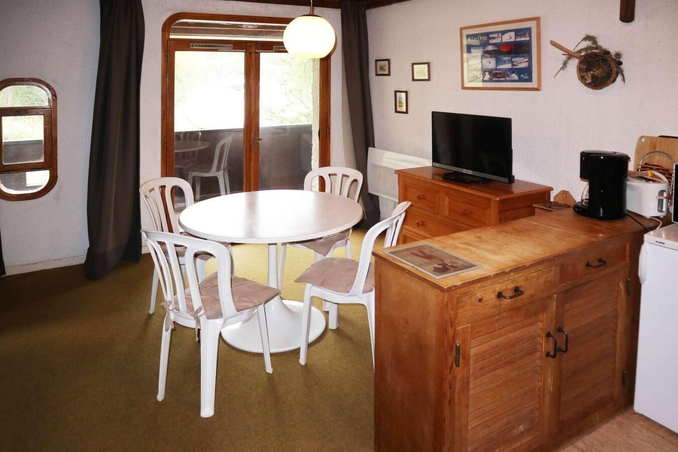 Ett kompakt kök med ett runt matbord, vita stolar, ett träskåp, en tv och en kaffebryggare.