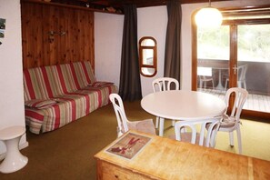 Ein gemütliches Zimmer mit einer gestreiften Couch, einem runden Tisch mit Stühlen, einer kleinen Holzkommode und einem Fenster mit Vorhängen.