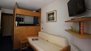 Een compacte kamer met een kitchenette, een bank, een televisie en een klein plankje met decoratieve objecten.