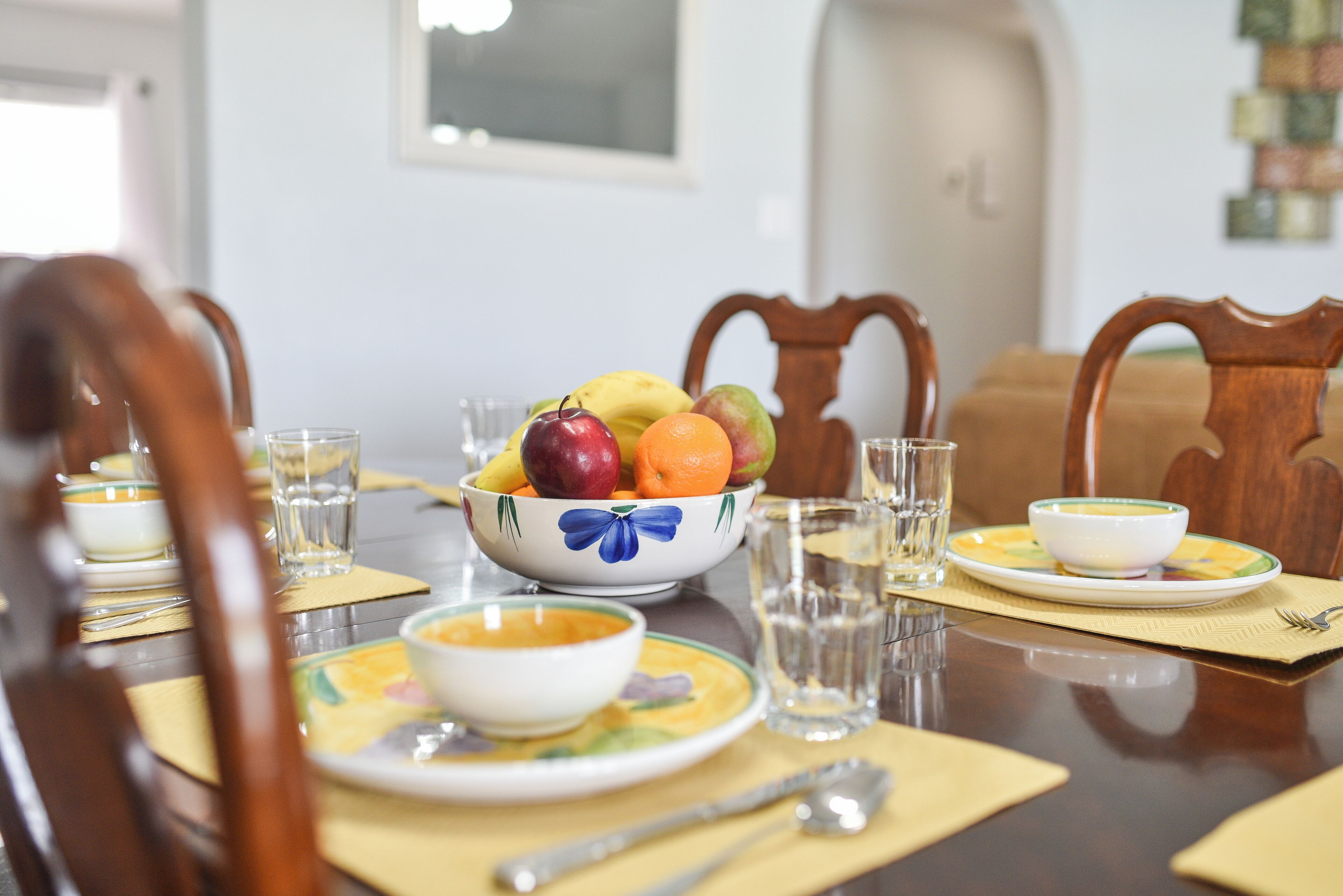 Una mesa de comedor lista con platos, tazones y vasos, y un frutero en el centro.