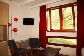 A room with a television mounted on the wall, two chairs, a bench, and a window with curtains.