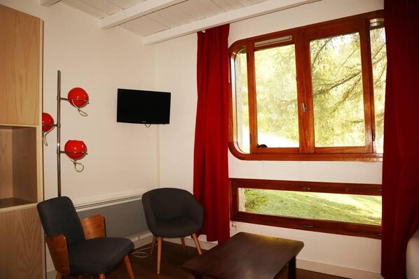 A room with a television mounted on the wall, two chairs, a bench, and a window with curtains.