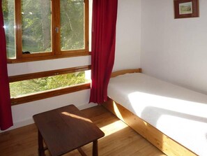 A small, single-room accommodation with a wooden bed, a small table, and a window with curtains.