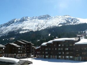 A snow-covered mountain with a clear sky. There are buildings with multiple floors and balconies, likely part of a hotel or resort. - Ski-in/ski-out & foot of the slopes, 1st floor, view ski slopes, terrace, tv, ski locker, 13m² (Valmorel)