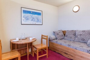 Ein gemütliches Zimmer mit einem Holztisch, Stühlen und einer Couch mit gemusterten Kissen. Ein gerahmtes Bild hängt an der Wand über dem Tisch.