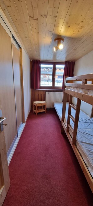 Ein Zimmer mit einem Etagenbett, einem kleinen hölzernen Hocker, einem Heizkörper und einem Fenster mit Vorhängen.