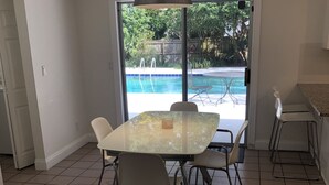 En matplats med ett glasbord och stolar, en hängande lampa och utsikt över en swimmingpool genom glasdörrar.