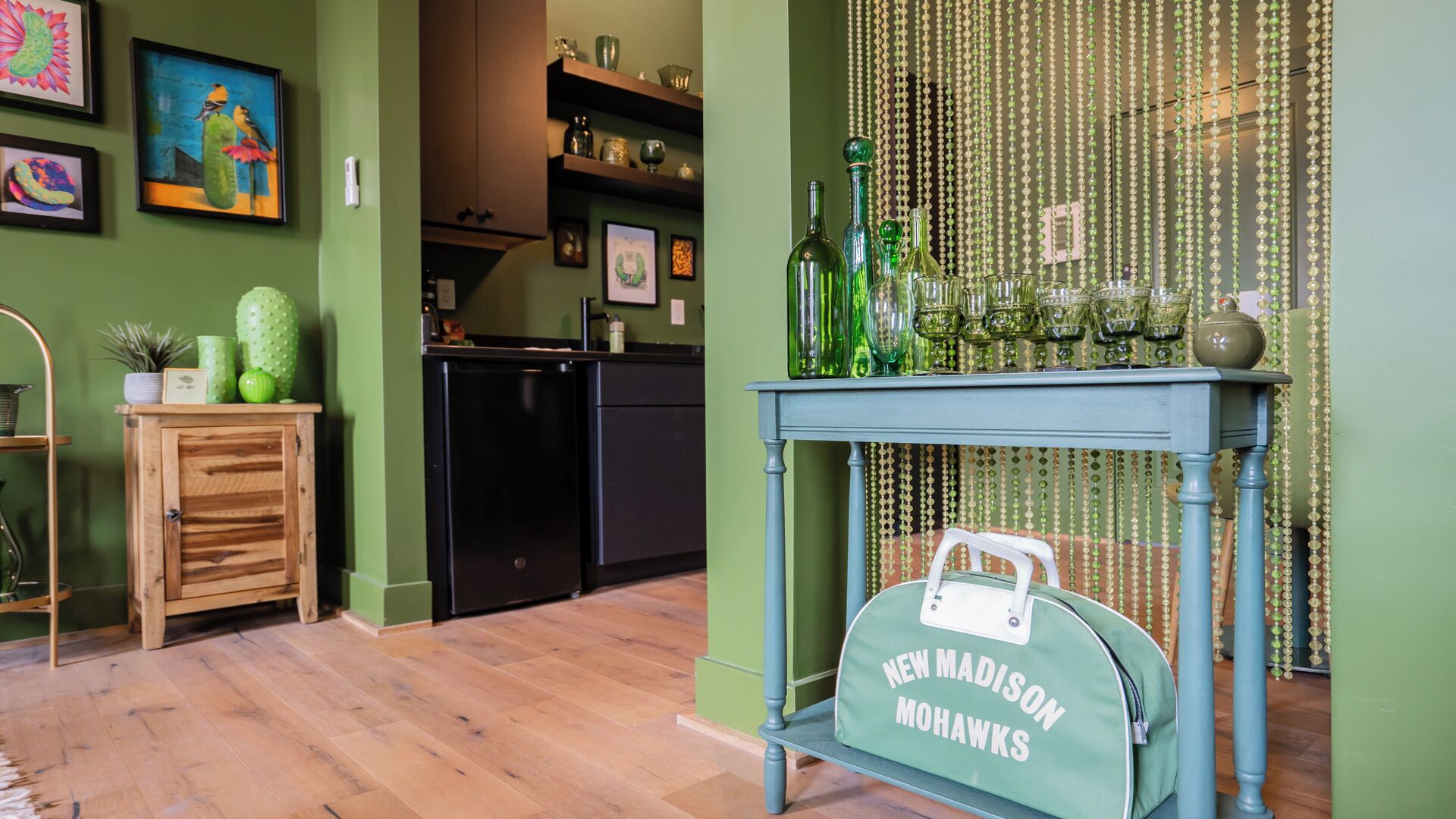 A kitchen with a wooden cabinet, a black refrigerator, and a blue table with a green bag labeled "NEW MADISON MOHAWKS".