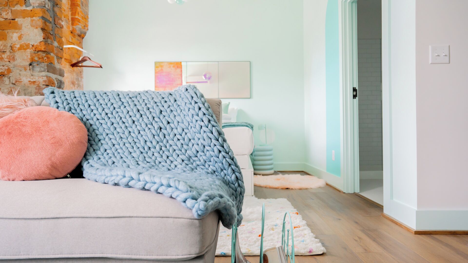 A cozy living room with a light-colored sofa, a chunky knit blanket, and a fluffy pillow. A teal-colored chair and a small table with a lamp are visible in the background.