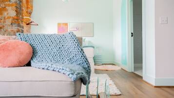 A cozy living room with a light-colored sofa, a chunky knit blanket, and a fluffy pillow. A teal-colored chair and a small table with a lamp are visible in the background.