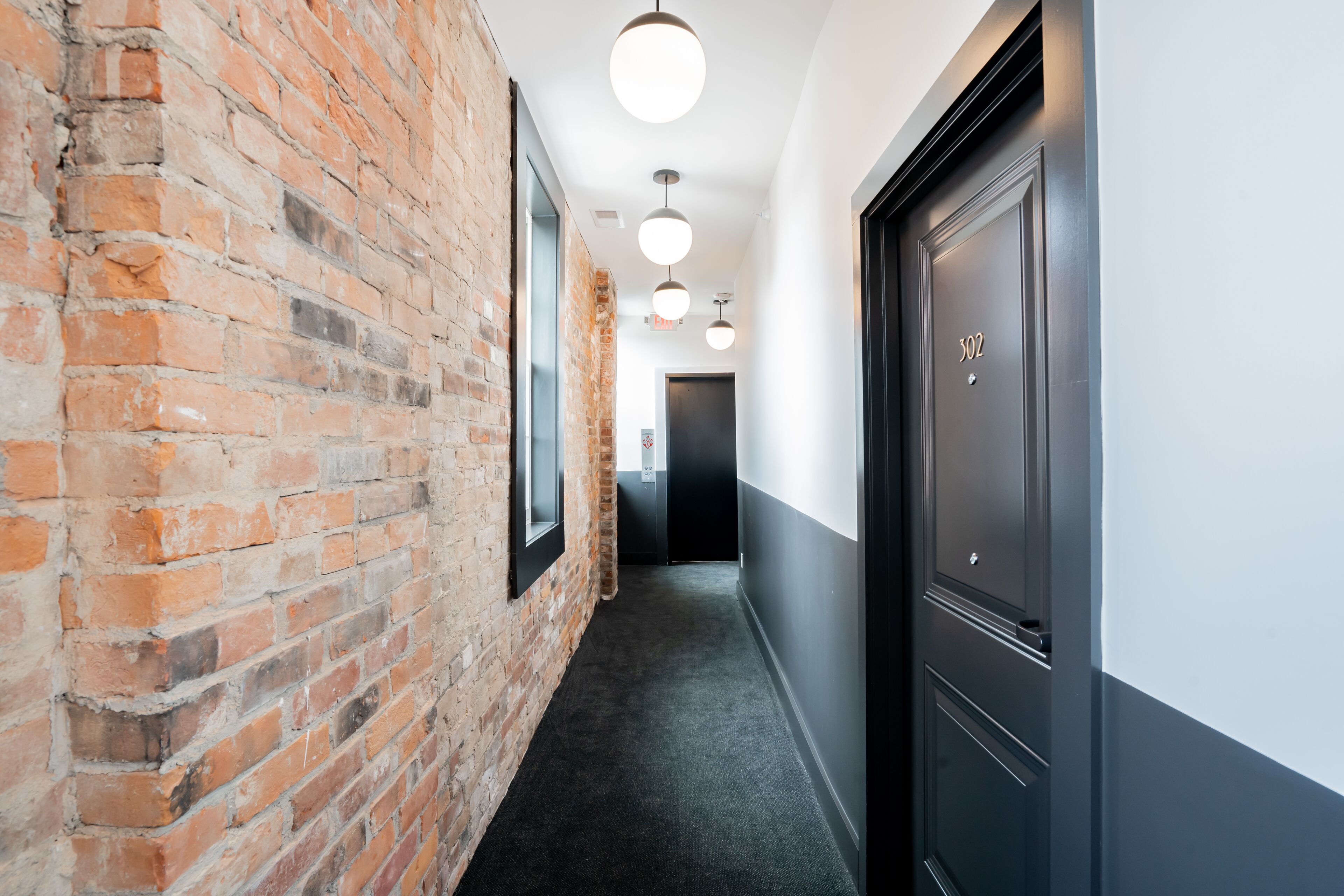 A hallway with a brick wall on one side and a dark door on the other. There are three ceiling lights and a framed picture on the brick wall.