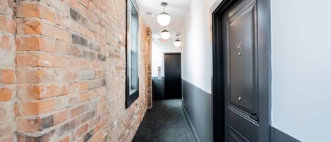 A hallway with a brick wall on one side and a dark door on the other. There are three ceiling lights and a framed picture on the brick wall.