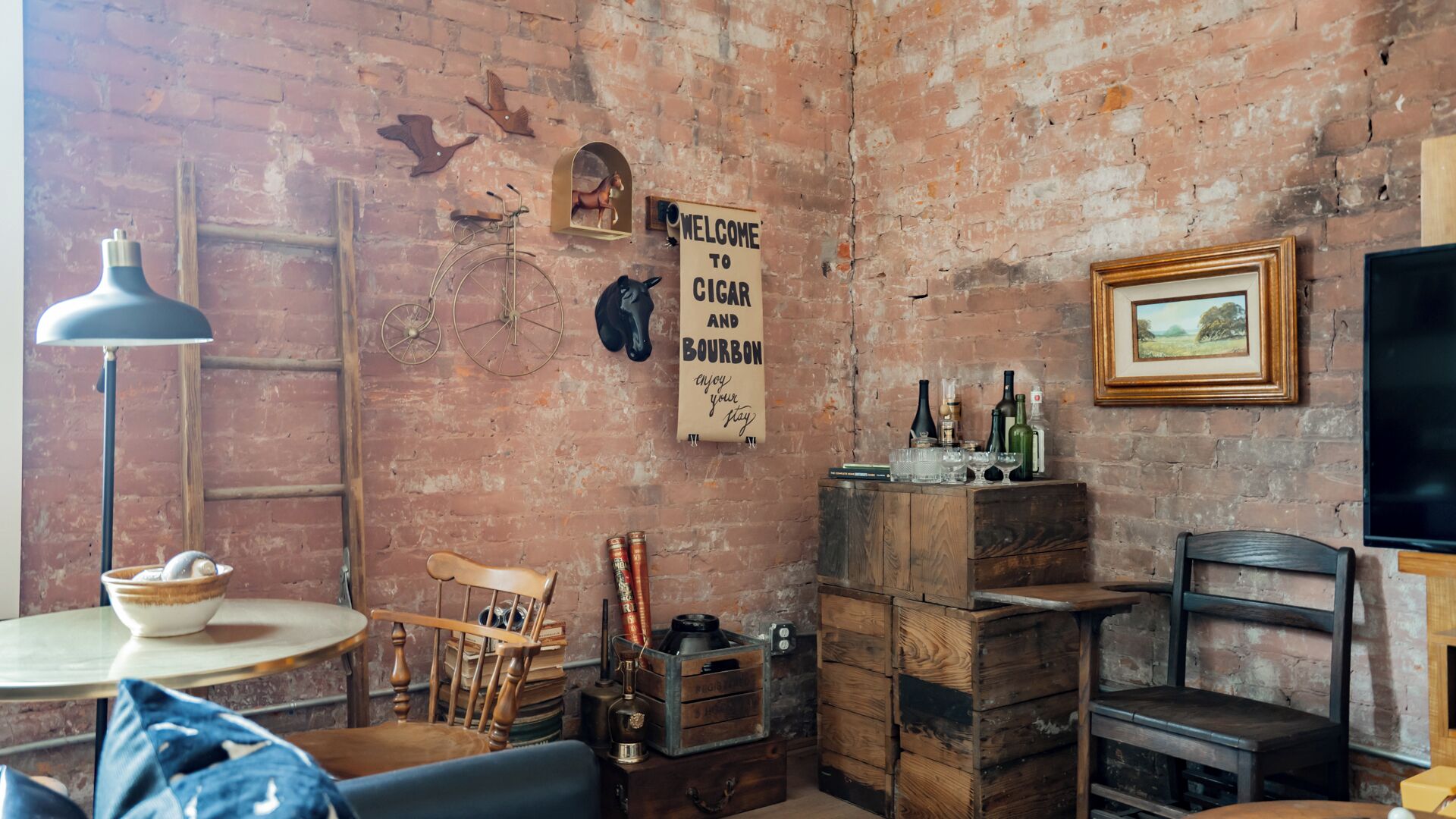 Una habitación rústica con paredes de ladrillo visto, una bicicleta antigua, una silla de madera, una pequeña mesa con una lámpara y un cartel dando la bienvenida a los visitantes para disfrutar de cigarros y bourbon.