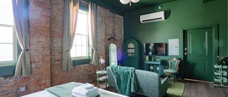 A room with a brick wall, a bed with a green blanket, a chair, a desk with a flat-screen TV, and an air conditioning unit.