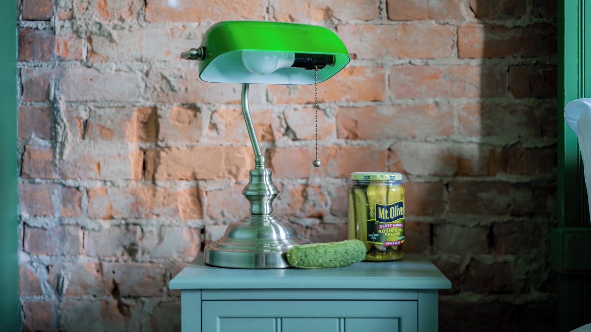 A vintage green desk lamp with a metal base and a jar of Mt. Olive Kosher dill spears on a white cabinet against a brick wall.
