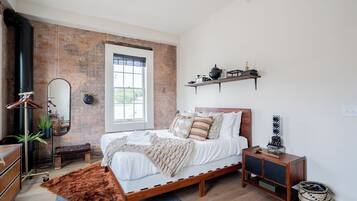 A bedroom with a brick wall, a large window, a bed with multiple pillows, a wooden bedside table, and a hanging shelf with decorative items.