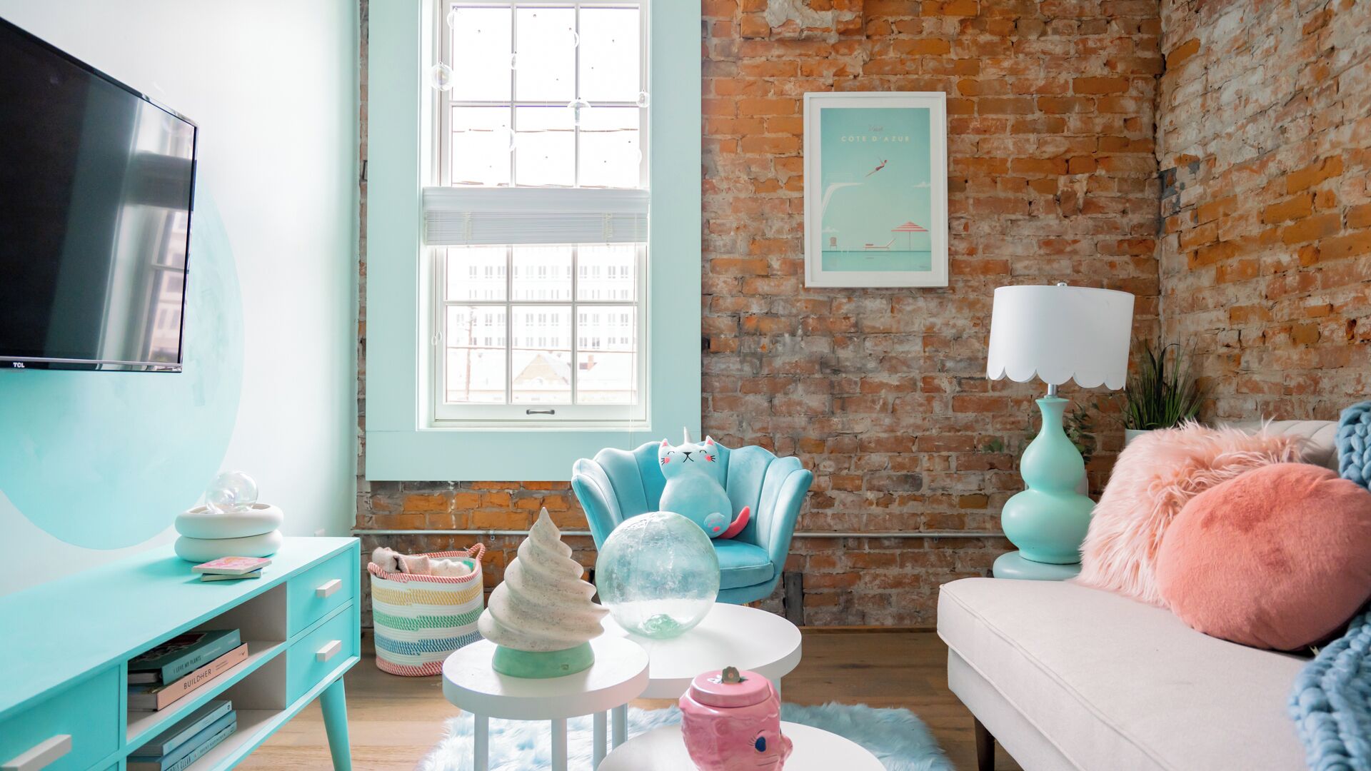 A cozy living room with a brick wall, a large window, a teal sofa, a white armchair, and a TV on the left.