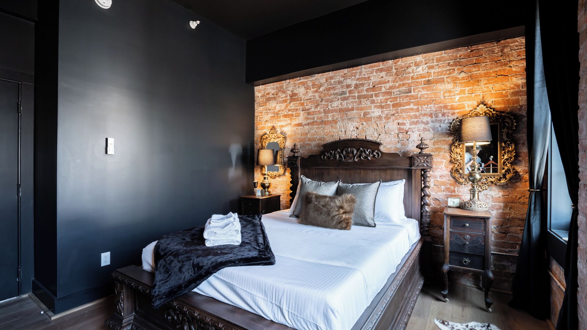 A modern bedroom with a brick wall, a wooden headboard, a large bed with white linens, two bedside tables with lamps, and a mirror on the wall.
