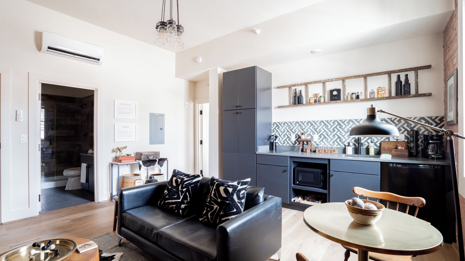 A modern living room with a black leather sofa, a dining table with a bowl, and a kitchen with a microwave and coffee maker.