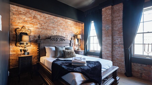 A bedroom with a brick wall, a large bed with multiple pillows, a nightstand, and a window with curtains.