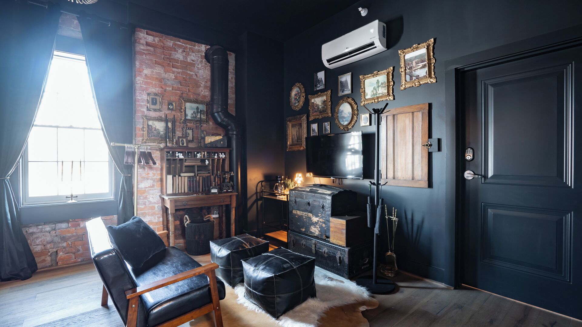 A room with a brick wall, a black leather chair, a vintage trunk, and a large black door.