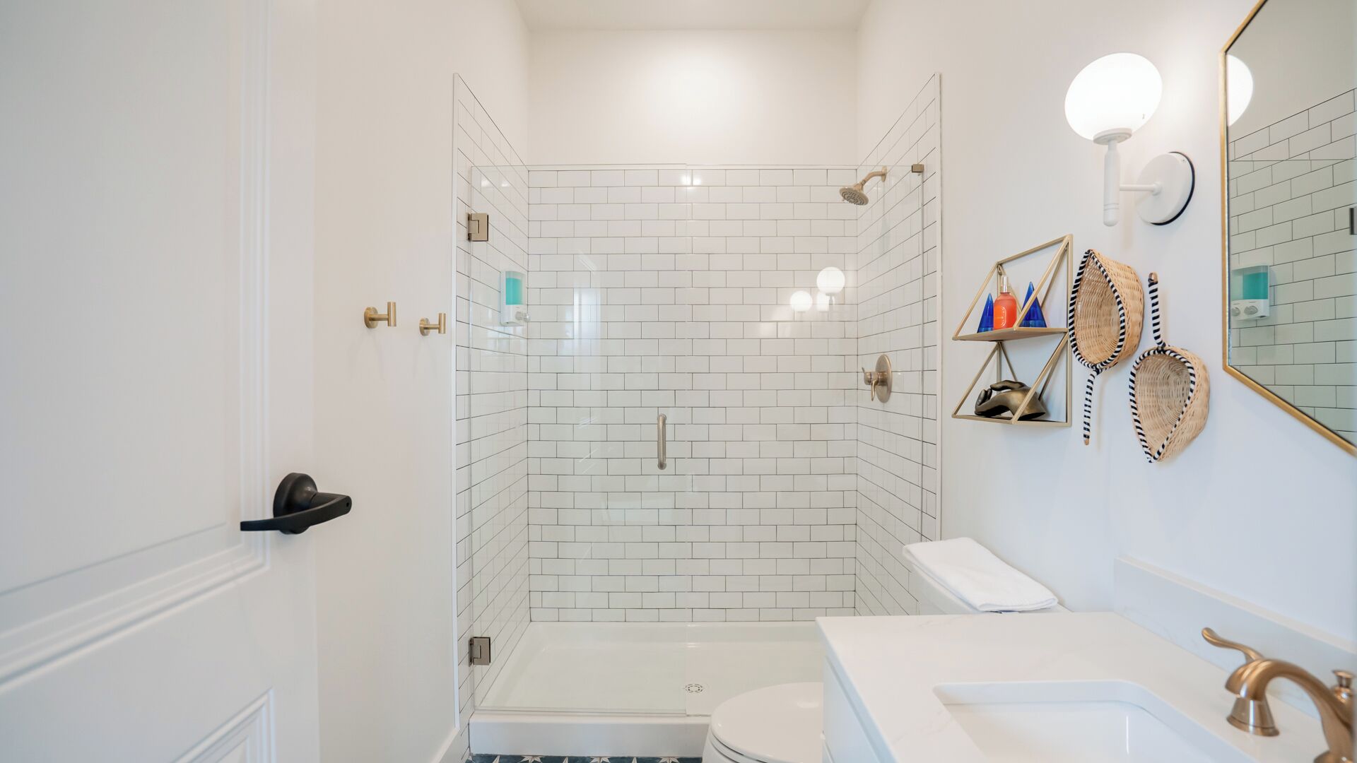 Un baño moderno con ducha de vidrio, paredes de azulejos blancos, inodoro blanco y lavamanos con grifo dorado.