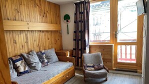 A wooden room with a sofa and a chair, a window with curtains, and a wall-mounted TV.