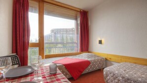 Habitación de hotel con dos camas, una mesa de comedor con mantel a cuadros, una ventana con cortinas y vistas a un edificio.